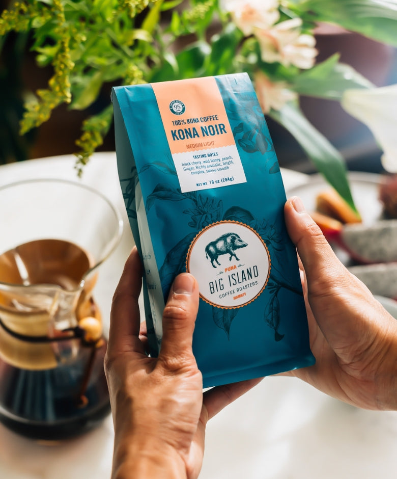 Person holding a blue coffee package labeled 'Kona Coffee Noir' by 'Big Island Coffee Roasters' with a blurred background of a kitchen setting.